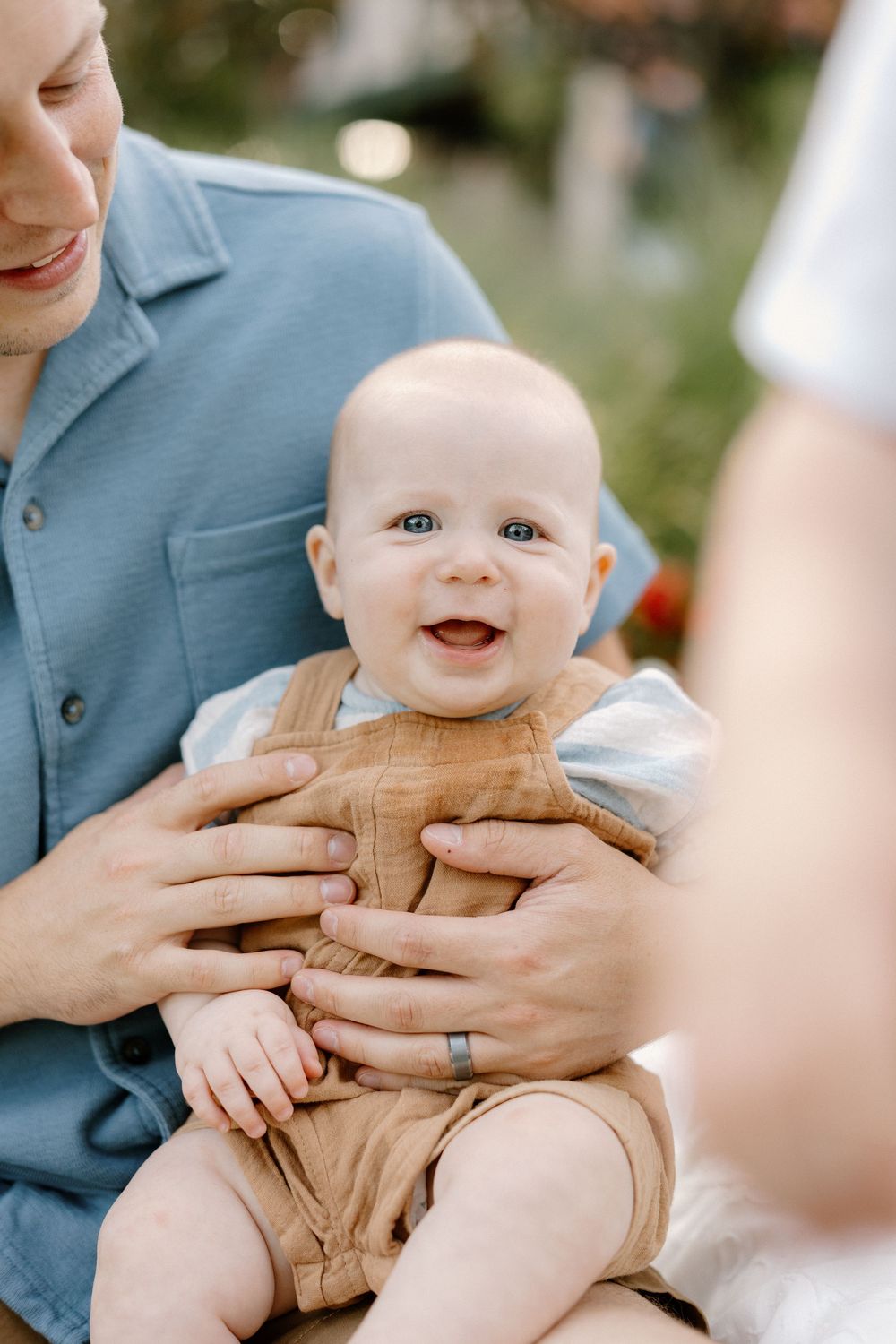 Sweet baby in a tan outfit smiles while being held against a soft outdoor background.