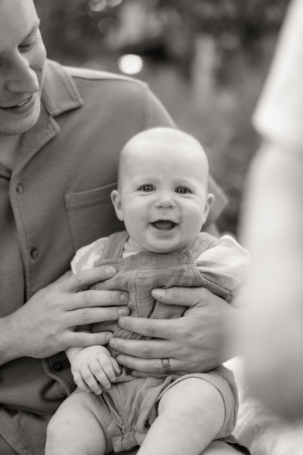A sweet baby wearing grey clothing shows a happy expression while being held in black and white photography.