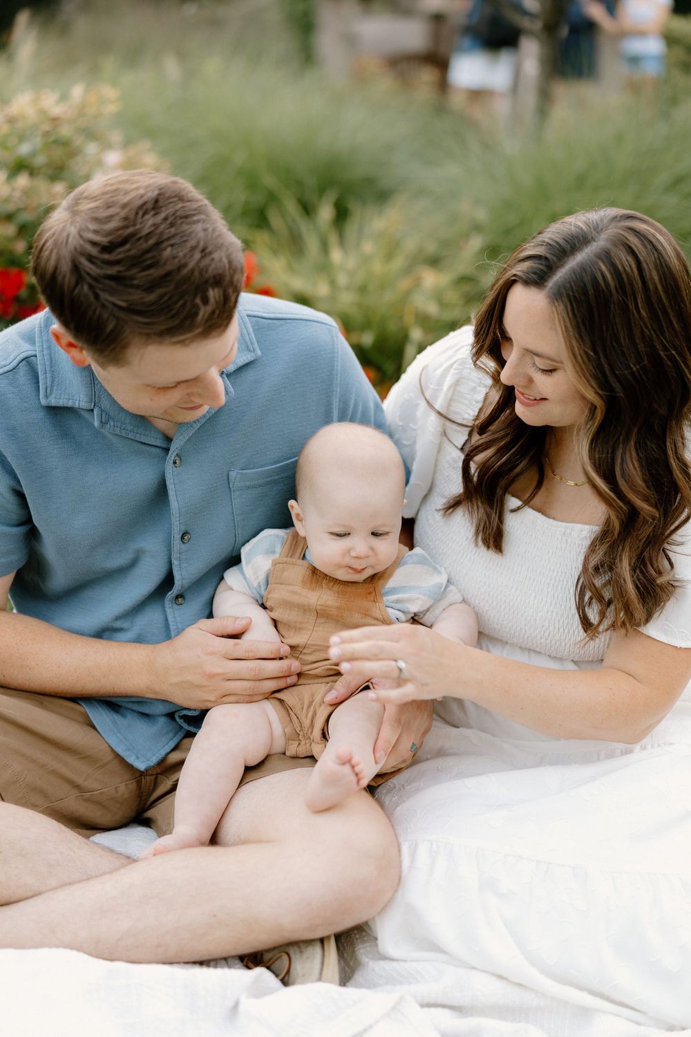 A loving couple lies on a white blanket outdoors, sharing tender moments with their newborn baby in matching blue and white outfits.
