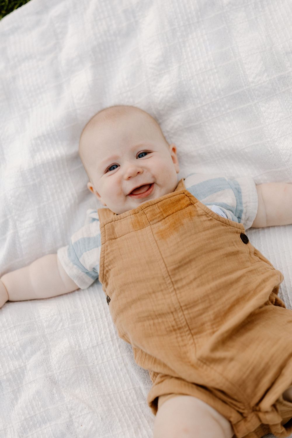 Happy baby wearing tan overalls and light blue shirt lying on white blanket.