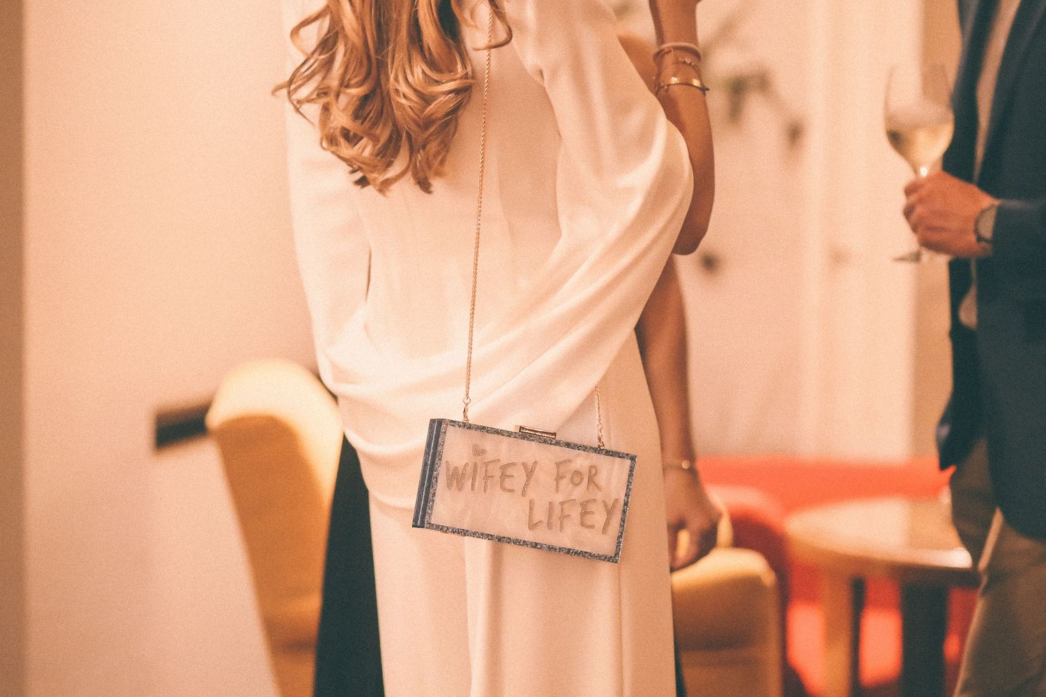 Person wearing white dress holds small purse in warm-toned indoor setting.