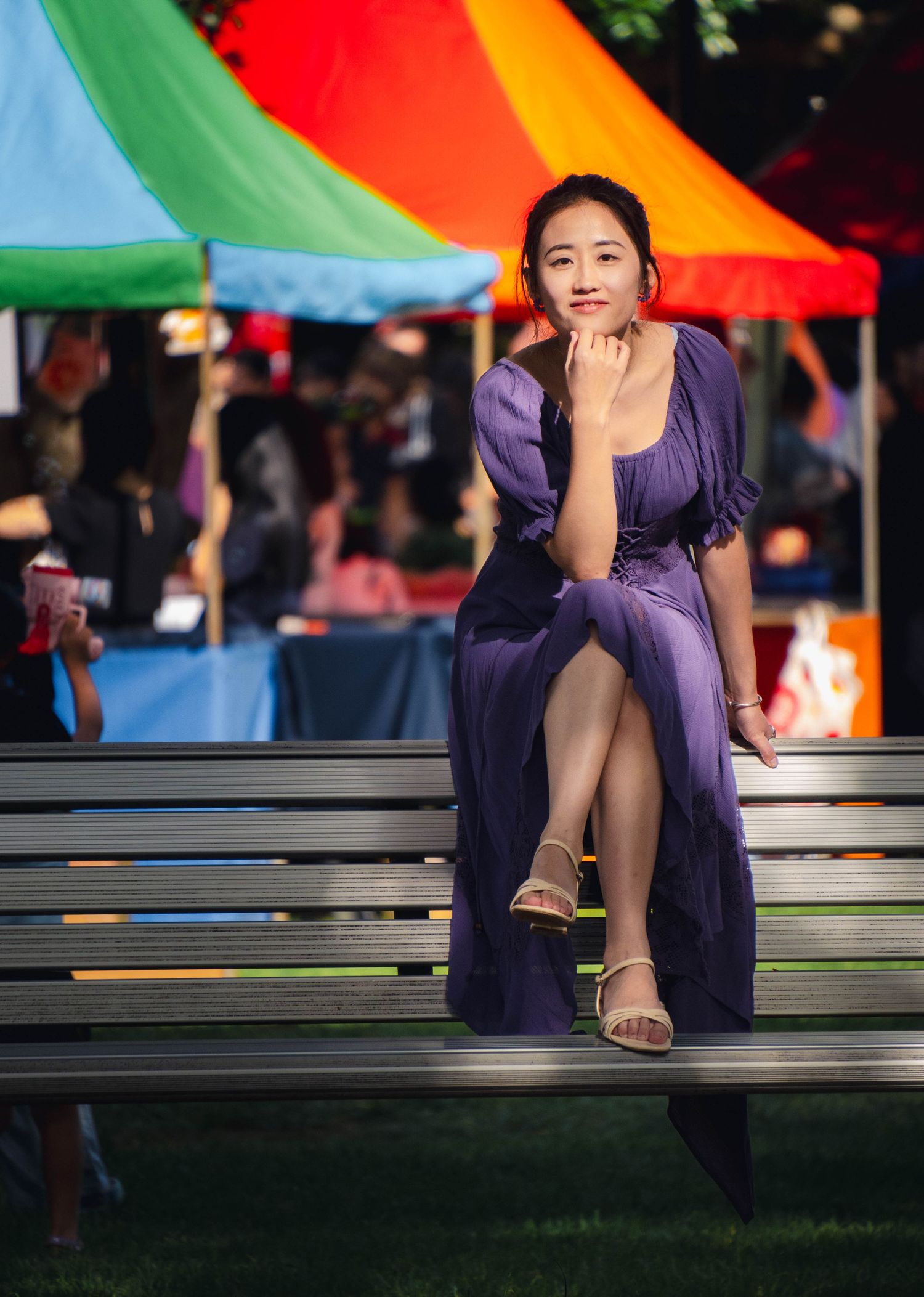 Asian lady in purple dress sitting on bench at colorful outdoor festival setting.