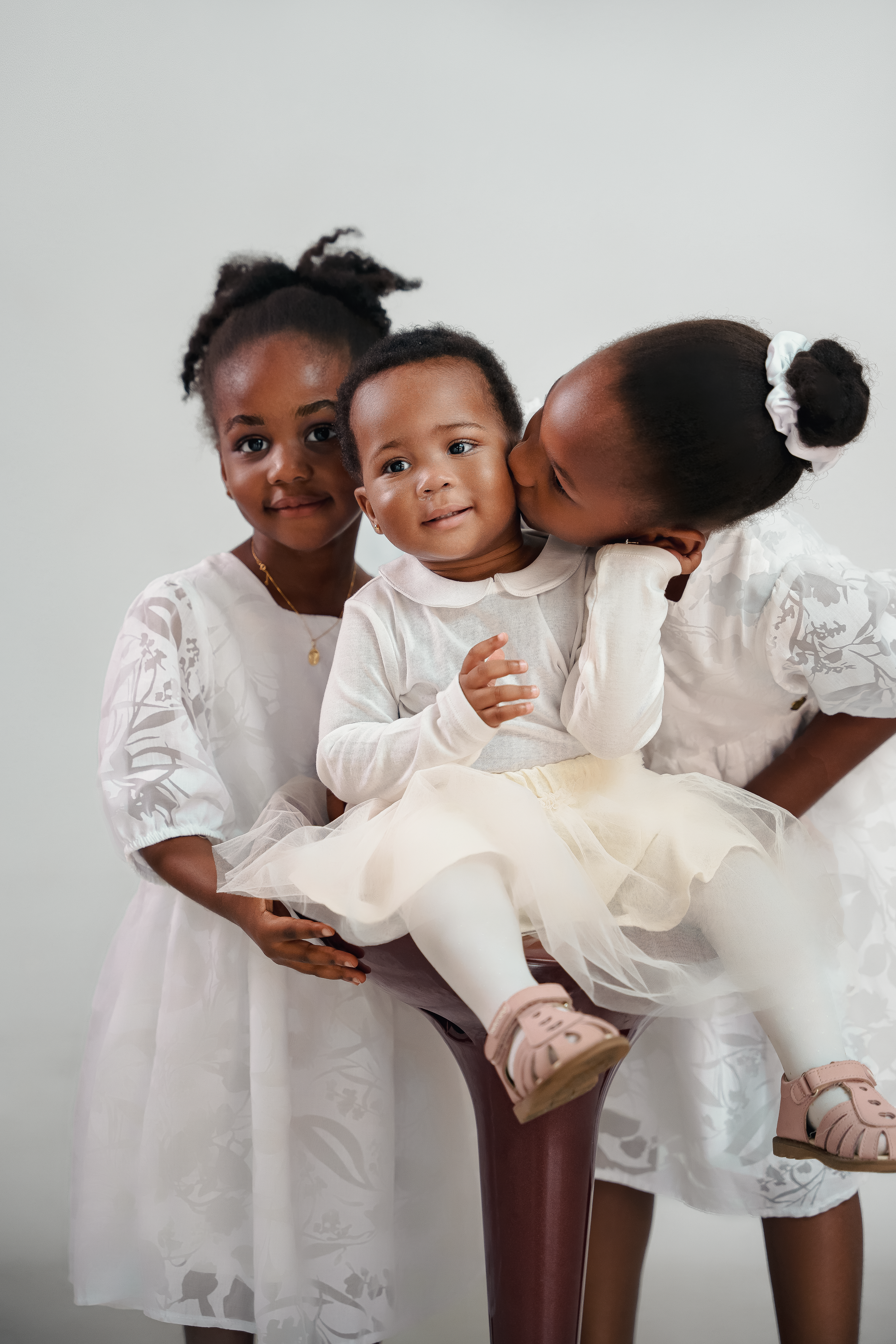 Three children in white dresses sharing a tender moment against a light background.