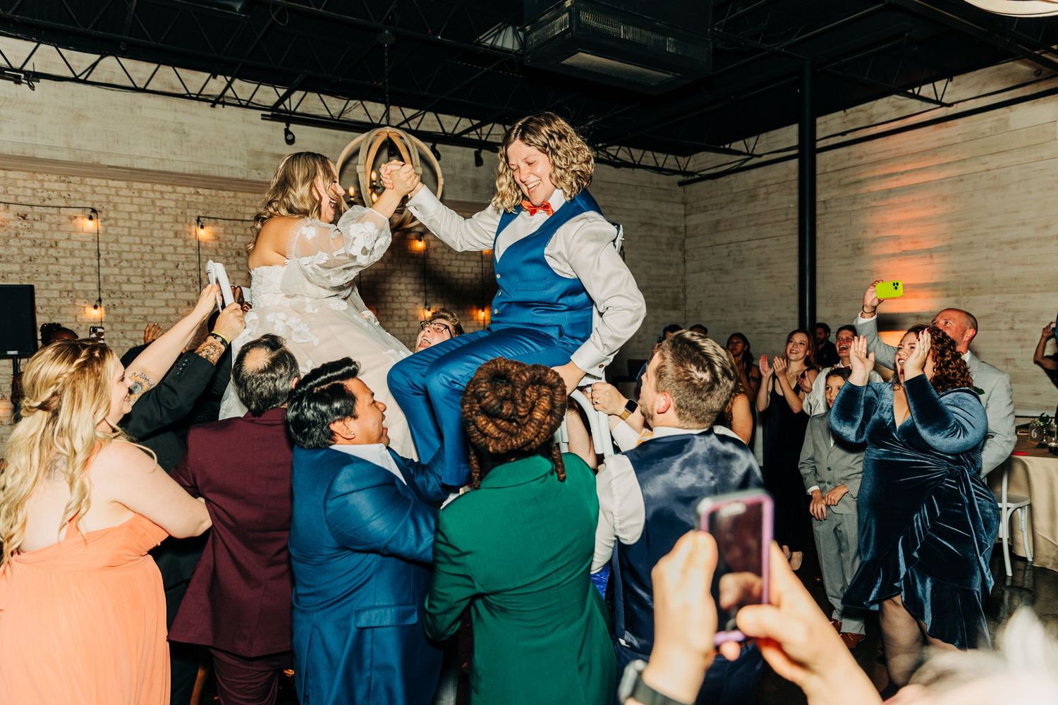 Wedding guests lift someone high in the air during celebratory dancing at a reception.
