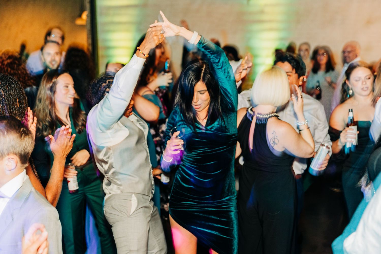 Wedding guests dance and celebrate on a dark dance floor with ambient lighting.