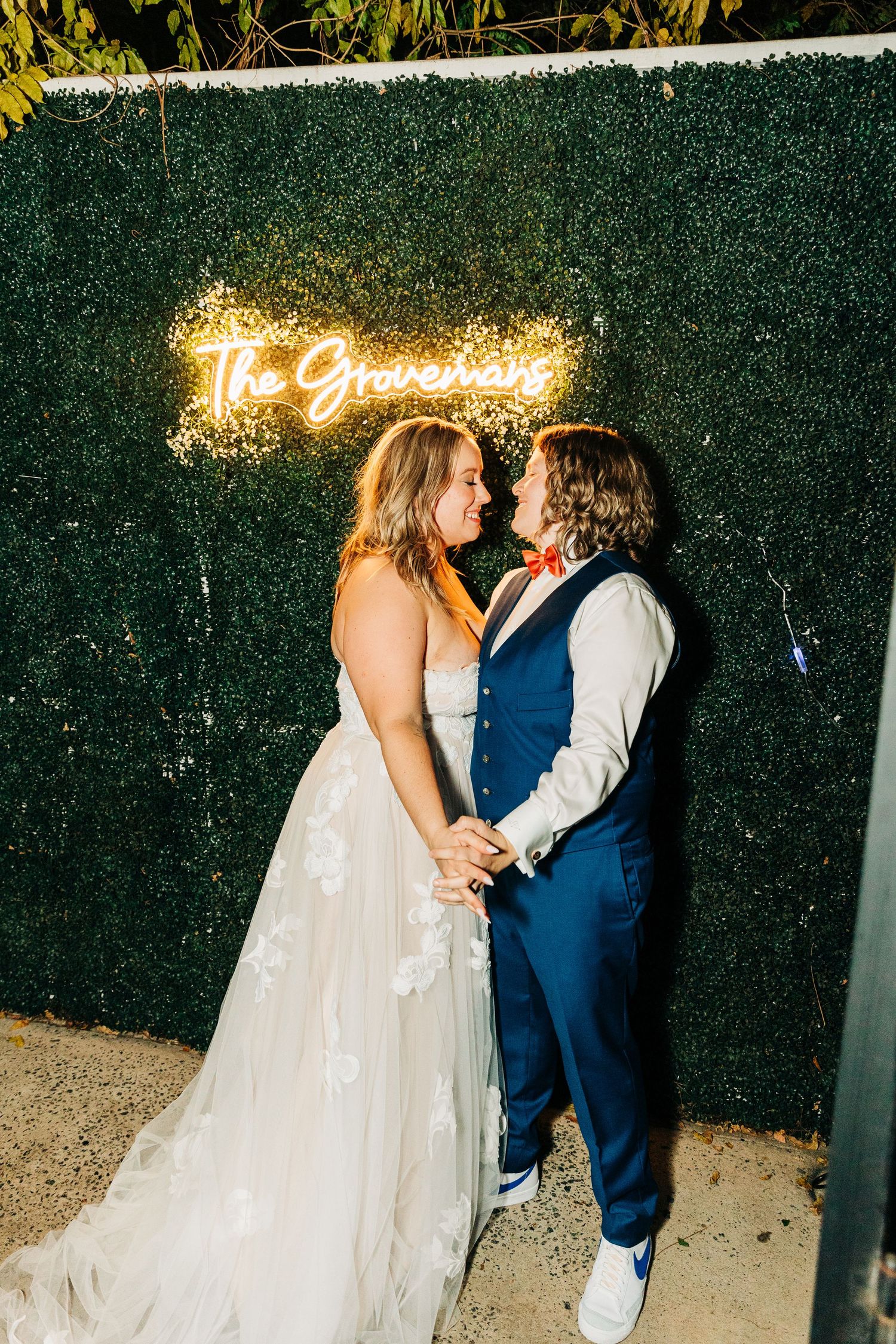 A couple poses against a green hedge wall with glowing neon signage.