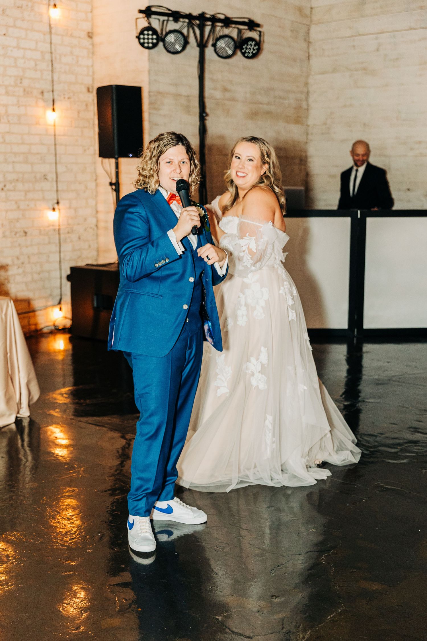 Two people share a moment at a wedding reception, one in a blue suit and sneakers and one in a white wedding gown.