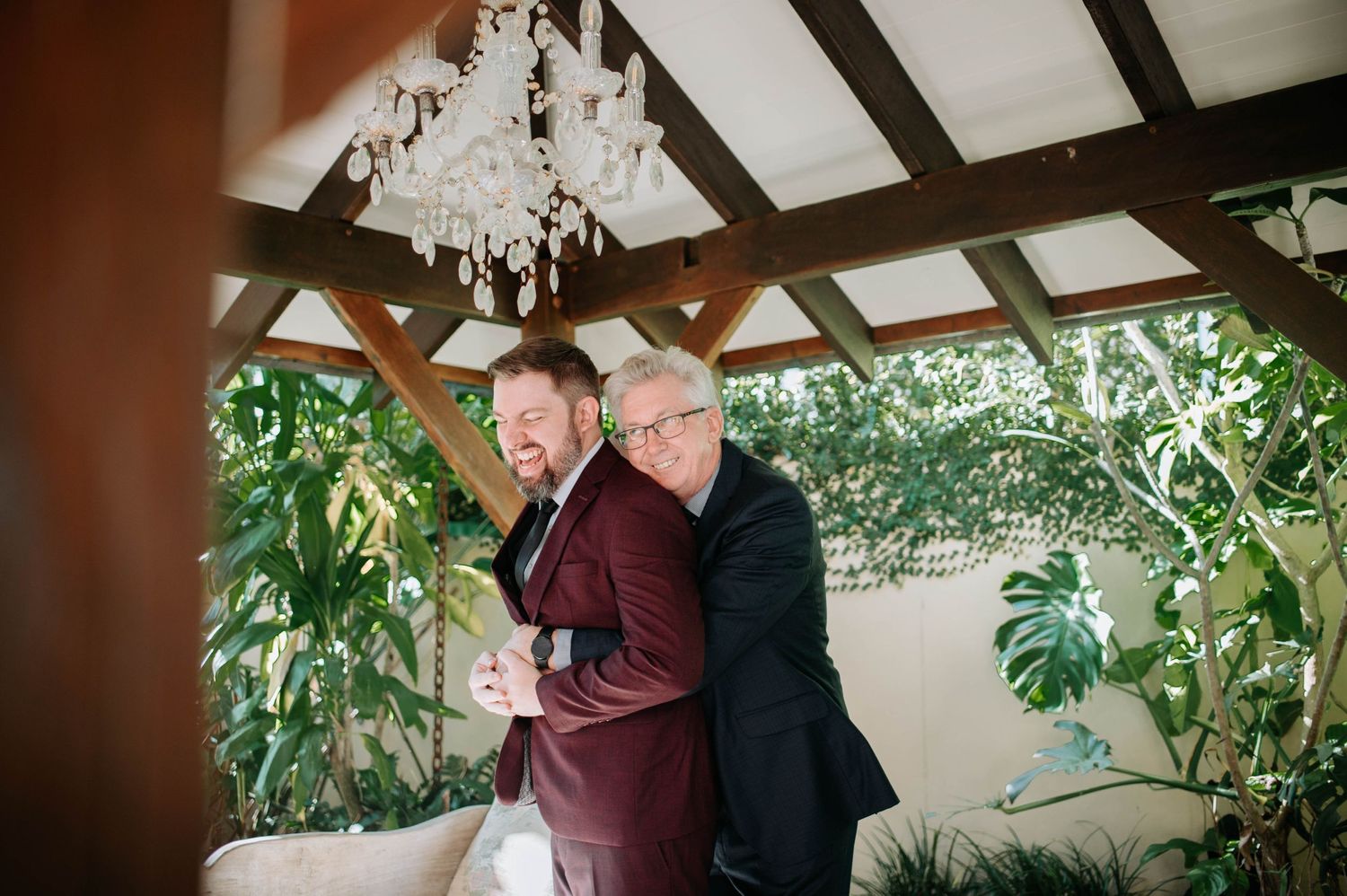 Two wedding guests share a playful moment under an elegant chandelier and tropical plants.
