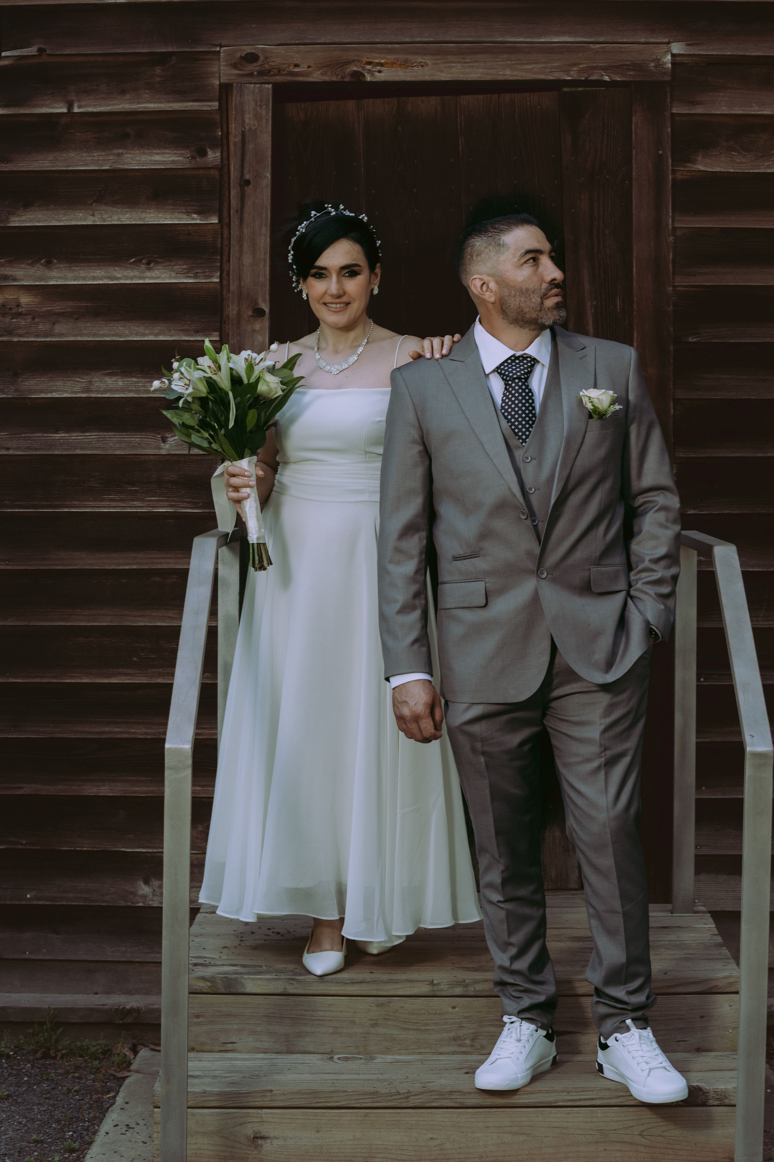 Wedding couple in white dress and grey suit standing by rustic wooden wall.