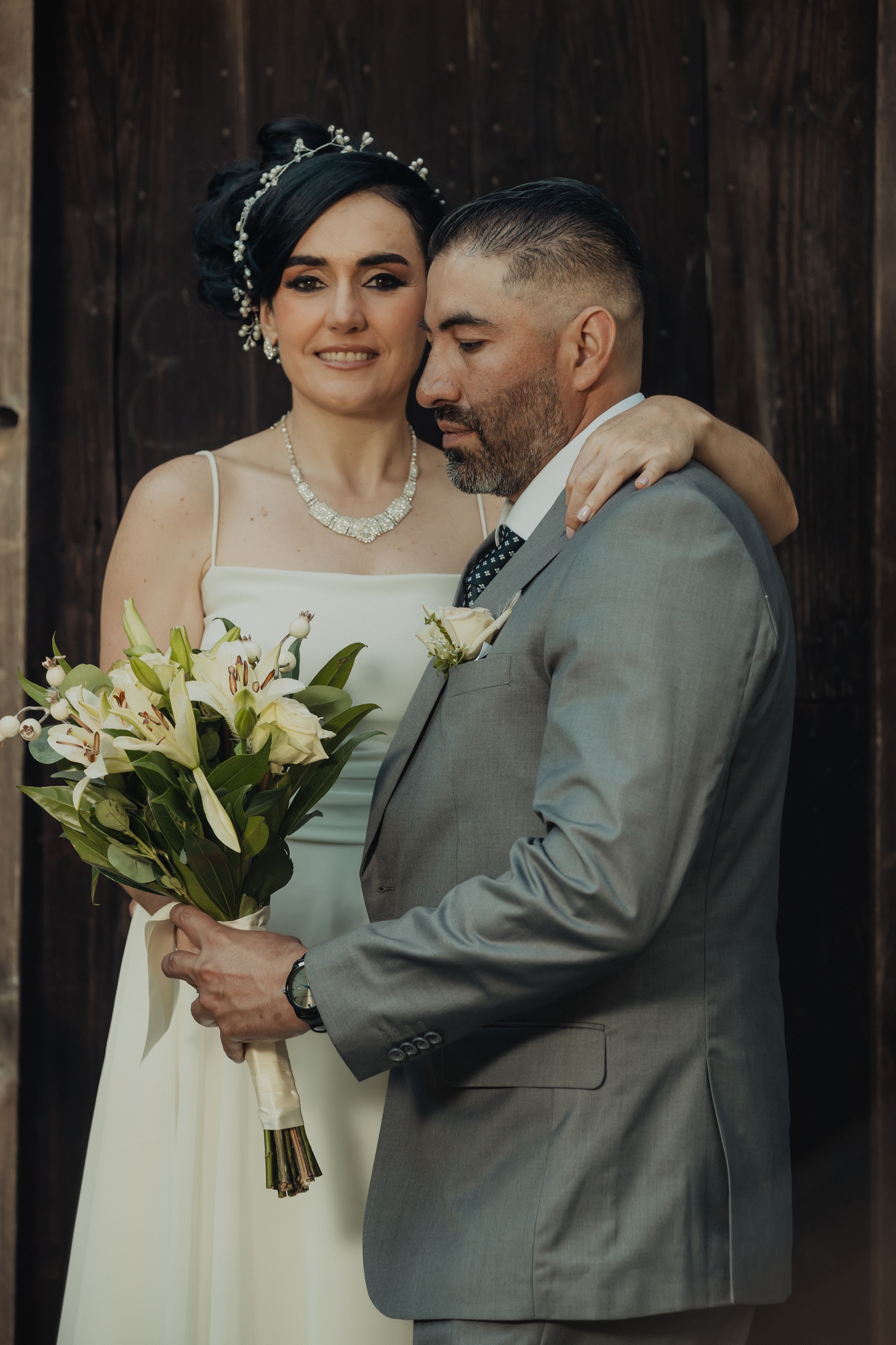 Bride in white dress holds bouquet while embracing groom in grey suit.