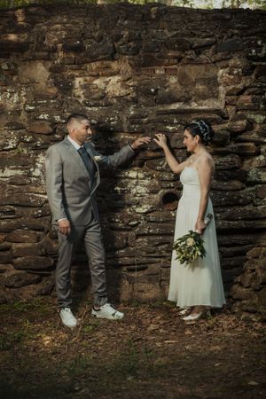 A couple in wedding attire poses playfully against a rustic stone wall during their outdoor photo session.
