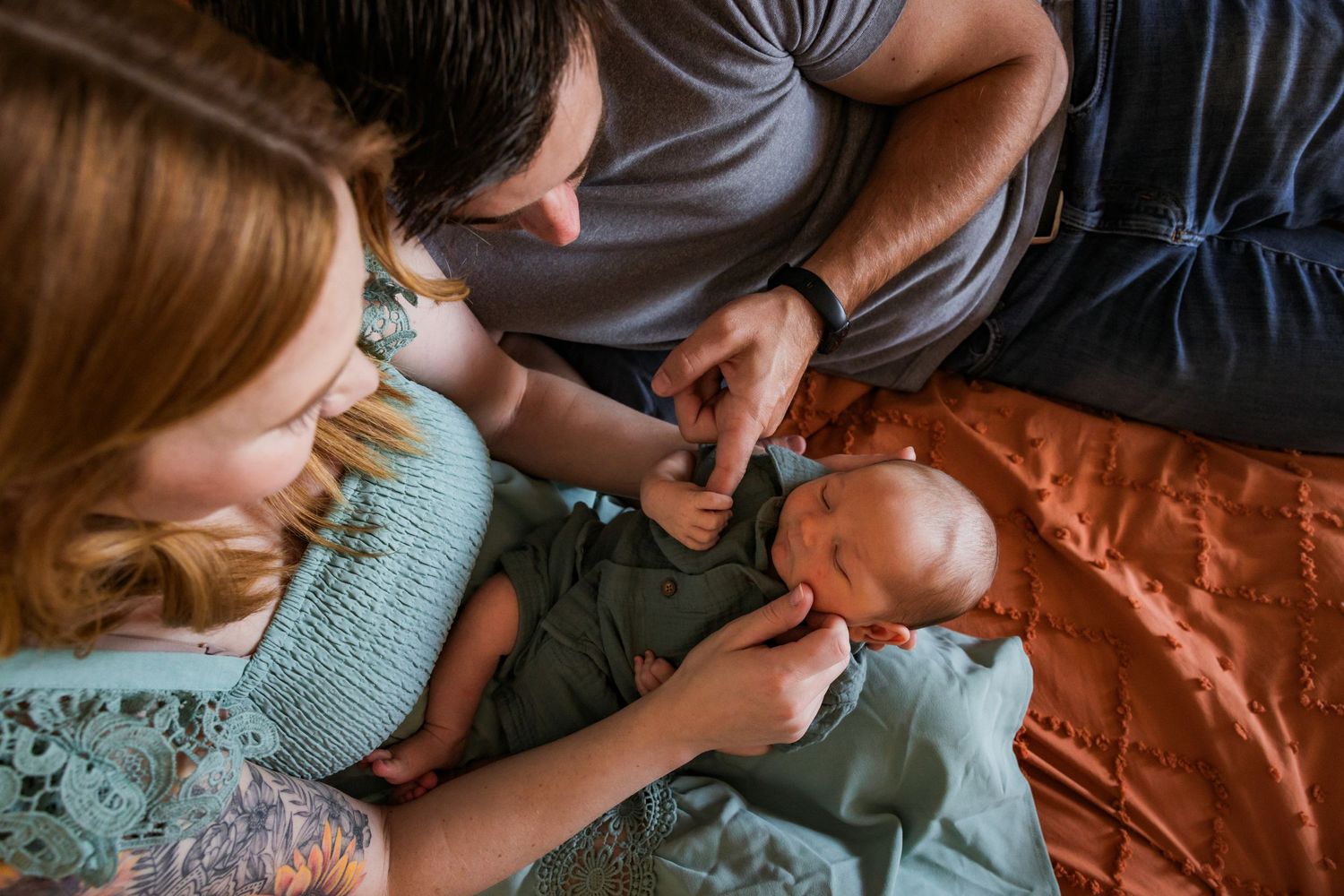 Father wearing a grey shirt holds a newborn hand while sitting with mother on an orange comforter.