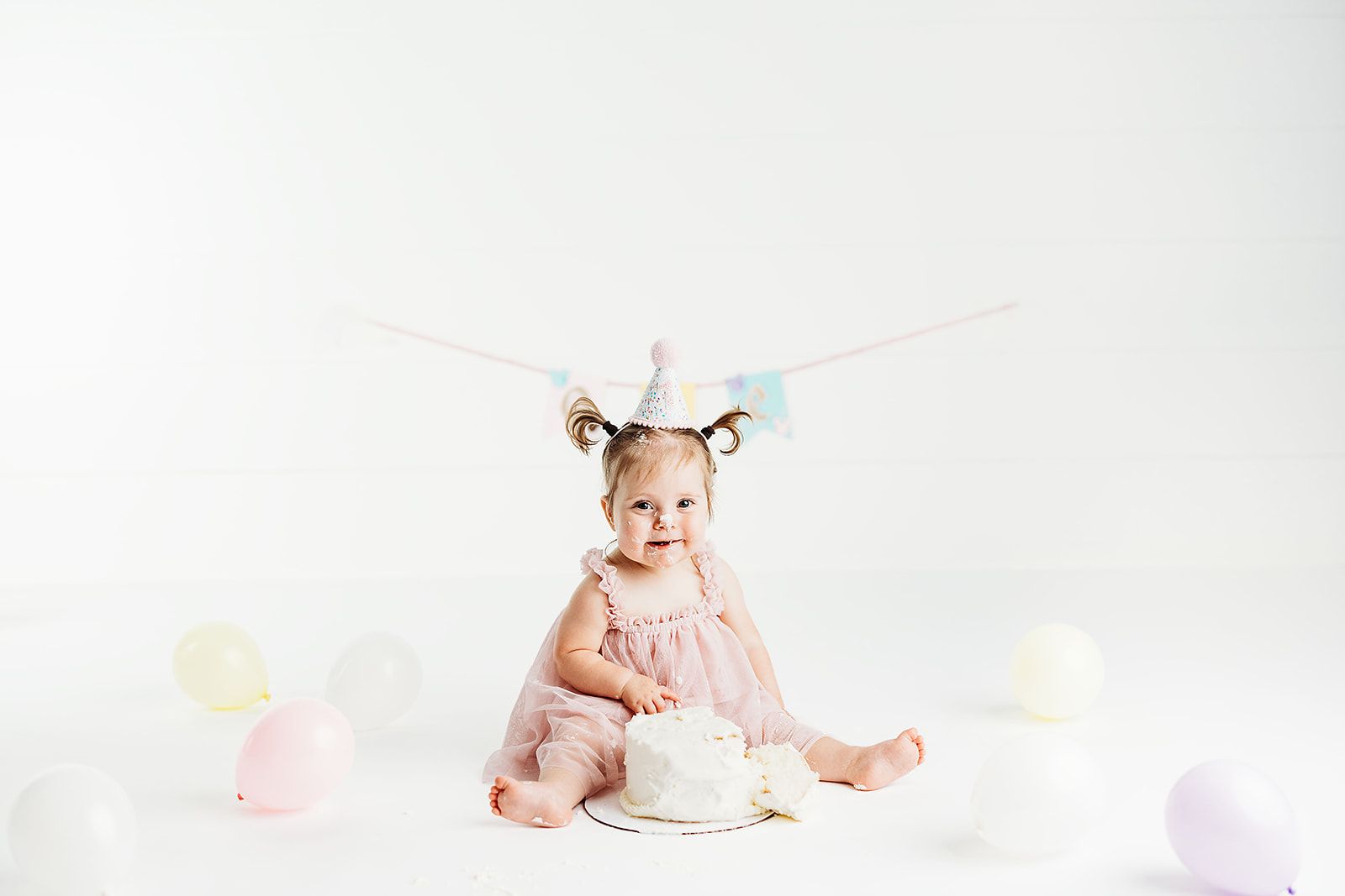 Toddler girl in pink dress and party hat sits with white smash cake and pastel balloons on white background.