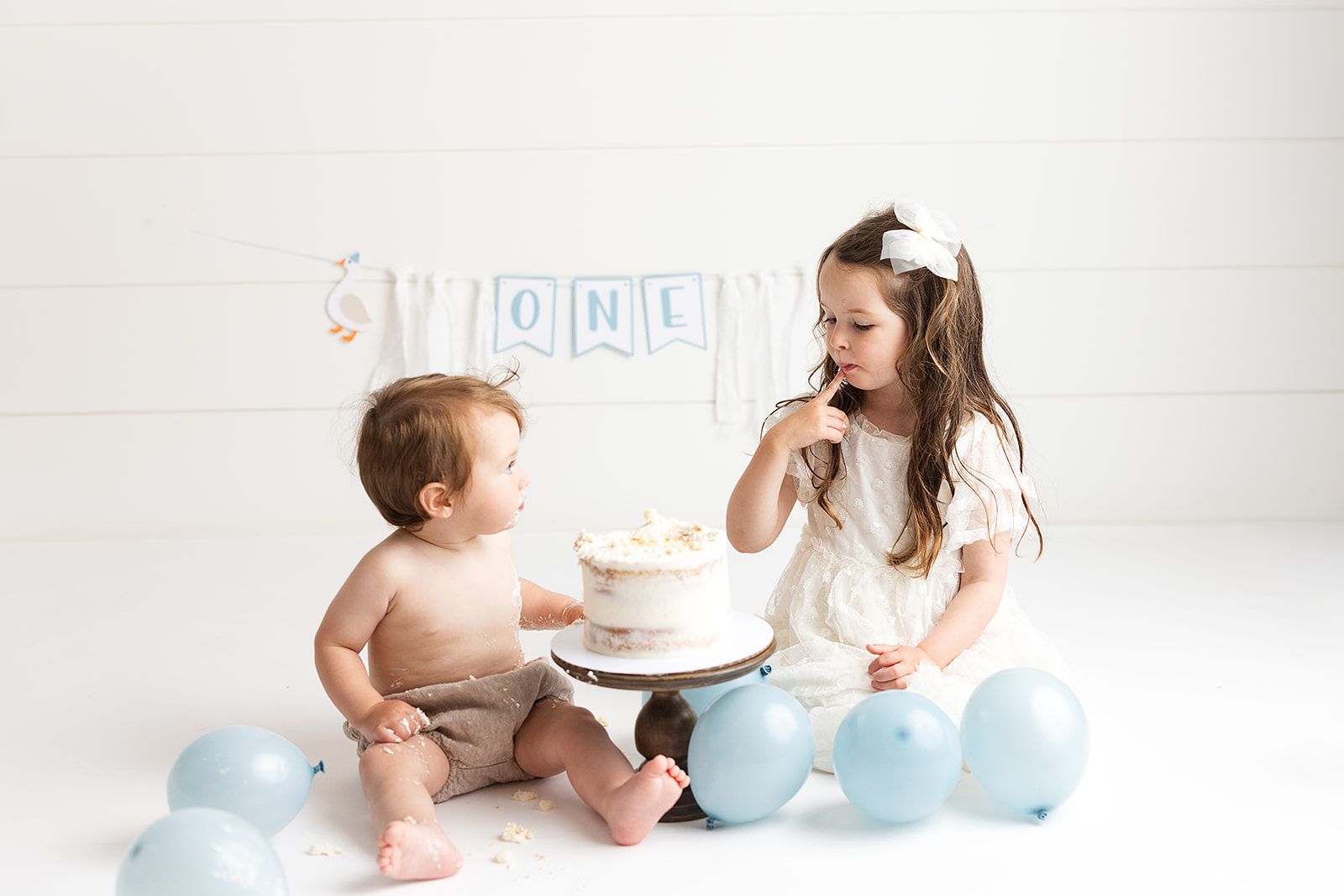 Baby's first birthday cake smash with older sister, blue balloons, and ONE banner on white background.