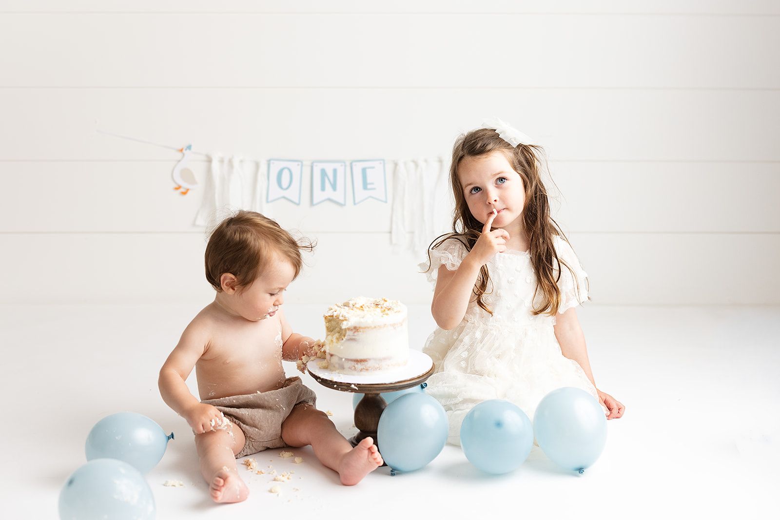 Baby's first birthday cake smash with older sister, blue balloons, and ONE banner on white background.