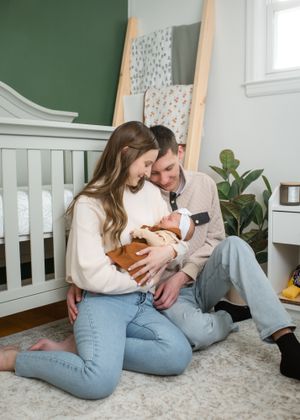 Parents sitting on nursery floor cuddling their newborn in a cozy, plant-filled room.