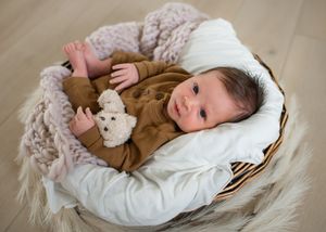 Baby lying in white basket with plush toy on cream blanket.