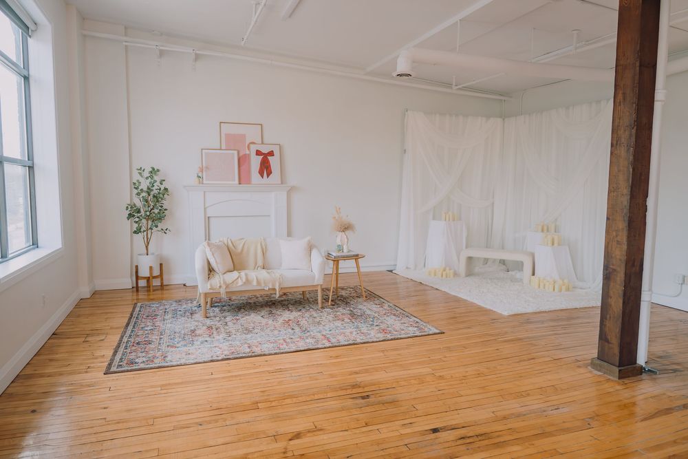 Bright minimalist studio space with hardwood floors, ornate rug, white furniture, and natural light from large windows.