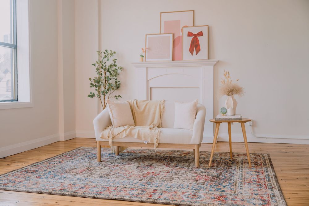 A cozy living room corner with a beige loveseat, vintage rug, potted plant, and pink wall art in a minimalist setting.