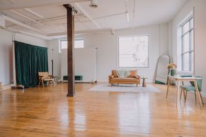 A bright studio space with wooden floors, emerald curtains, and scattered furniture including a tan sofa and side tables.