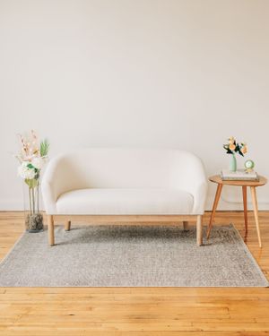 A white curved sofa with wooden legs on a gray rug, accompanied by a small wooden side table.