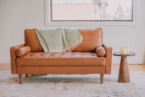 Mid-century modern tan leather sofa with sage green throw blanket and wooden side table in minimalist living room.