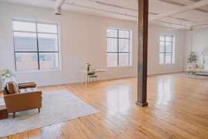 Bright open loft space with hardwood floors, white walls, exposed ceiling beams and large windows letting in natural light.