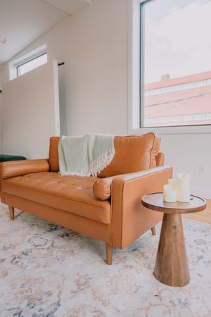 Modern tan leather sofa with tufted cushions sits on floral patterned rug beside wooden pedestal side table in bright room.