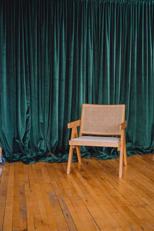 Three wooden chairs with cane seats stand in a row against a dark teal velvet curtain on a hardwood floor.