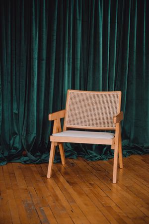 A wooden bench with cane webbing seat back is positioned against a dark green curtain on a hardwood floor.