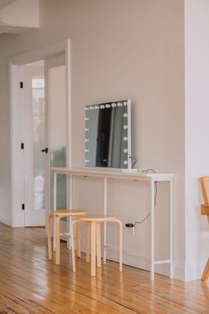 Modern vanity setup with illuminated mirror, white console table and wooden stools on hardwood floors in minimal room.