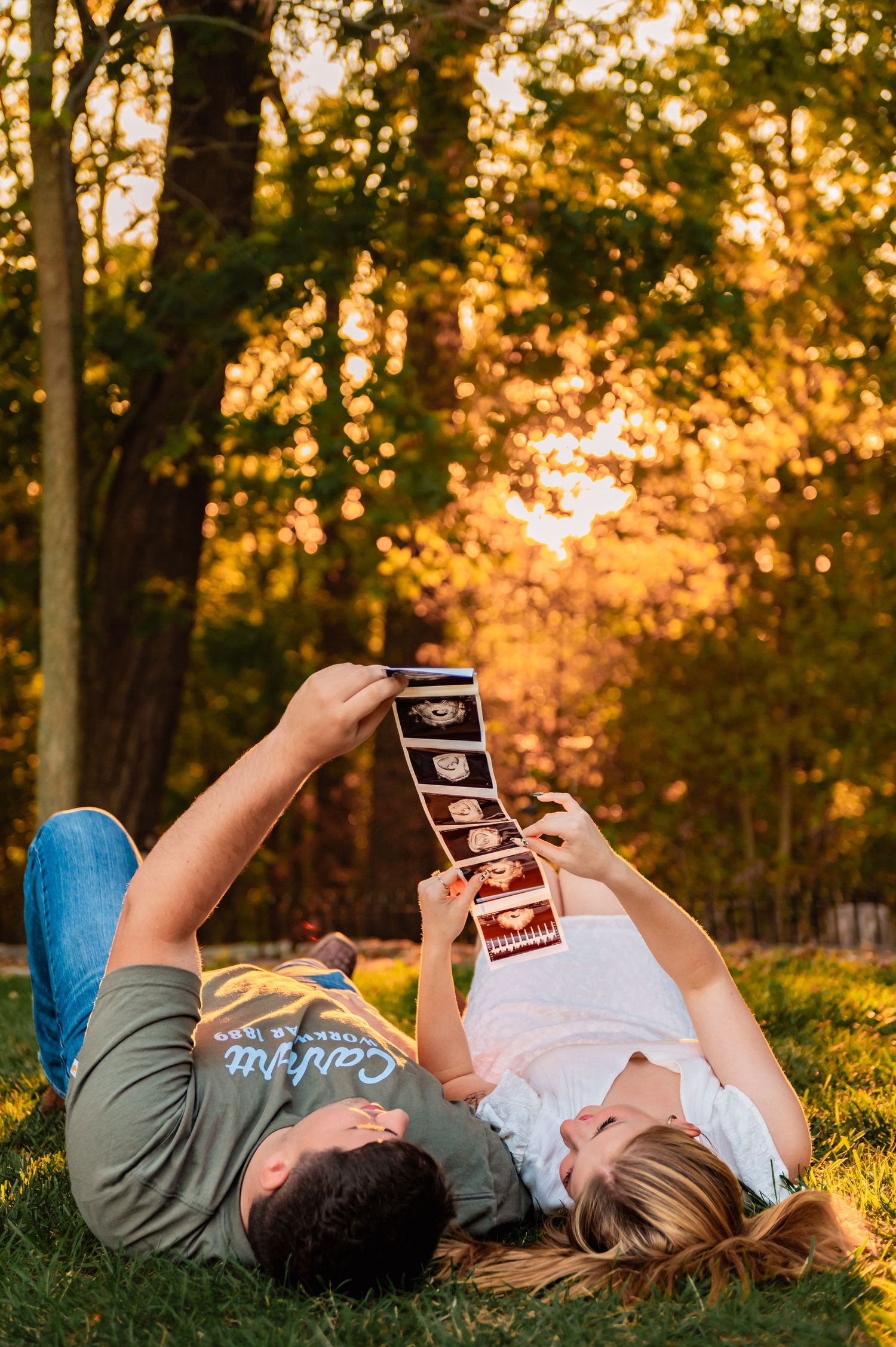 Couple lying on autumn ground looking at ultrasound photos in golden sunset light.
