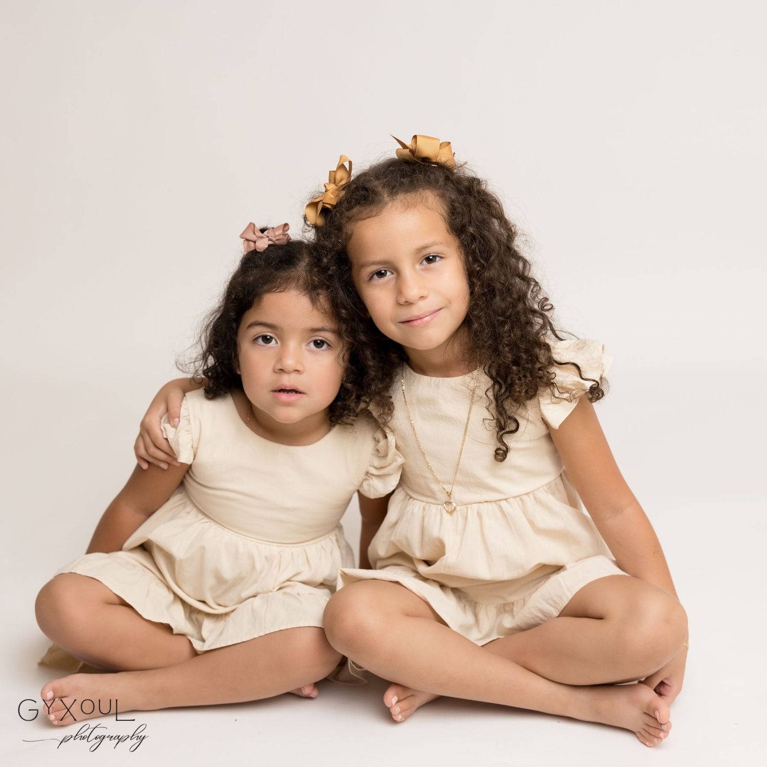 Two children in matching cream dresses sitting together for a portrait.