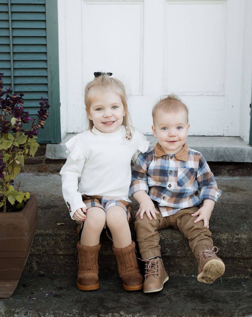 Two young children sitting on stone steps in front of a white door with green shutters.
