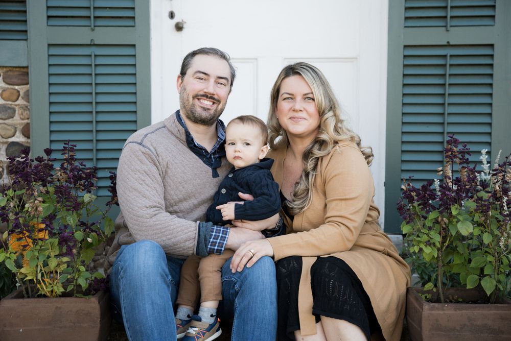 A family of three poses together on a wooden bench with flowering plants in the background.