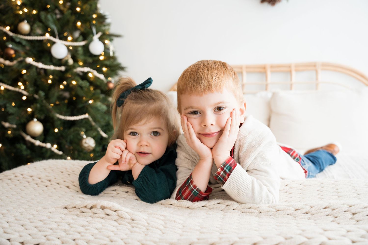 Two children laying on white blanket pose playfully in front of decorated Christmas tree.