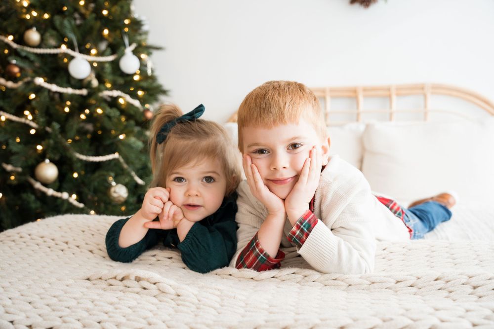 Two children laying on white blanket pose playfully in front of decorated Christmas tree.