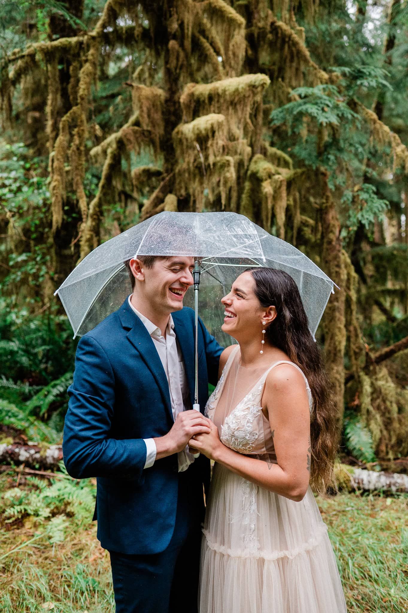 bride and groom laughing while holding clear umbrella in mossy rainforest in Washington State