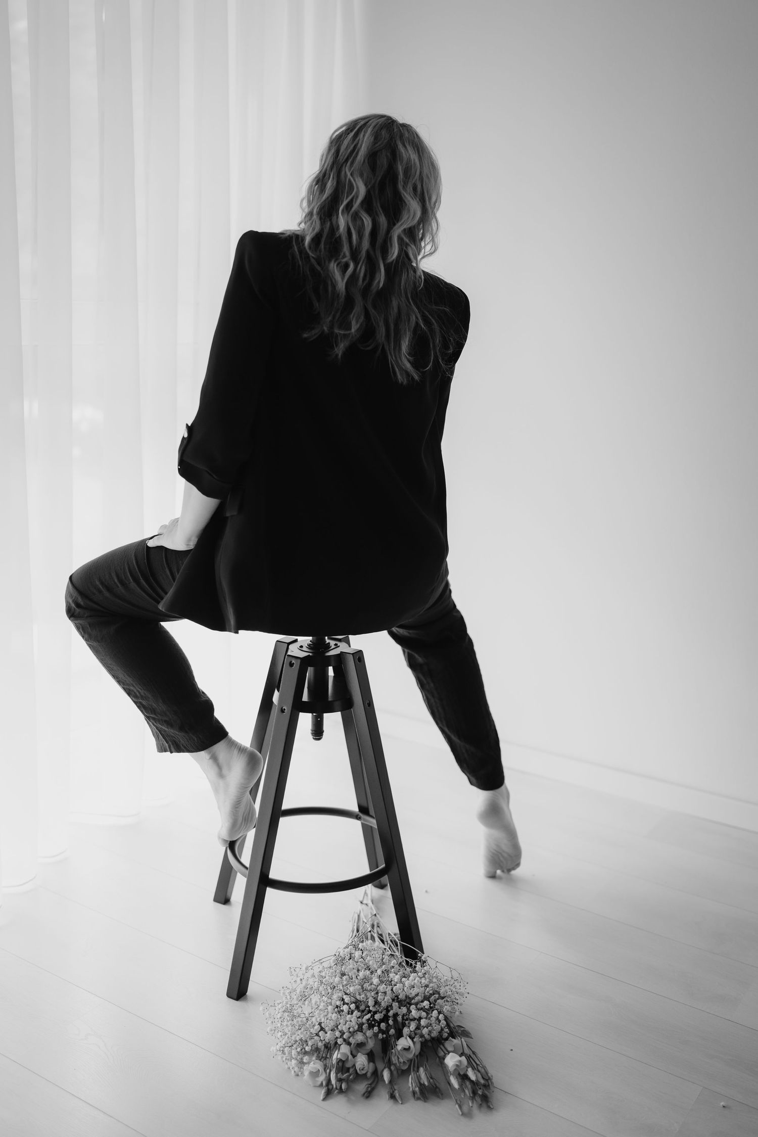 Black and white artistic photo of someone in dark clothing sitting backwards on stool with flowers on floor below.