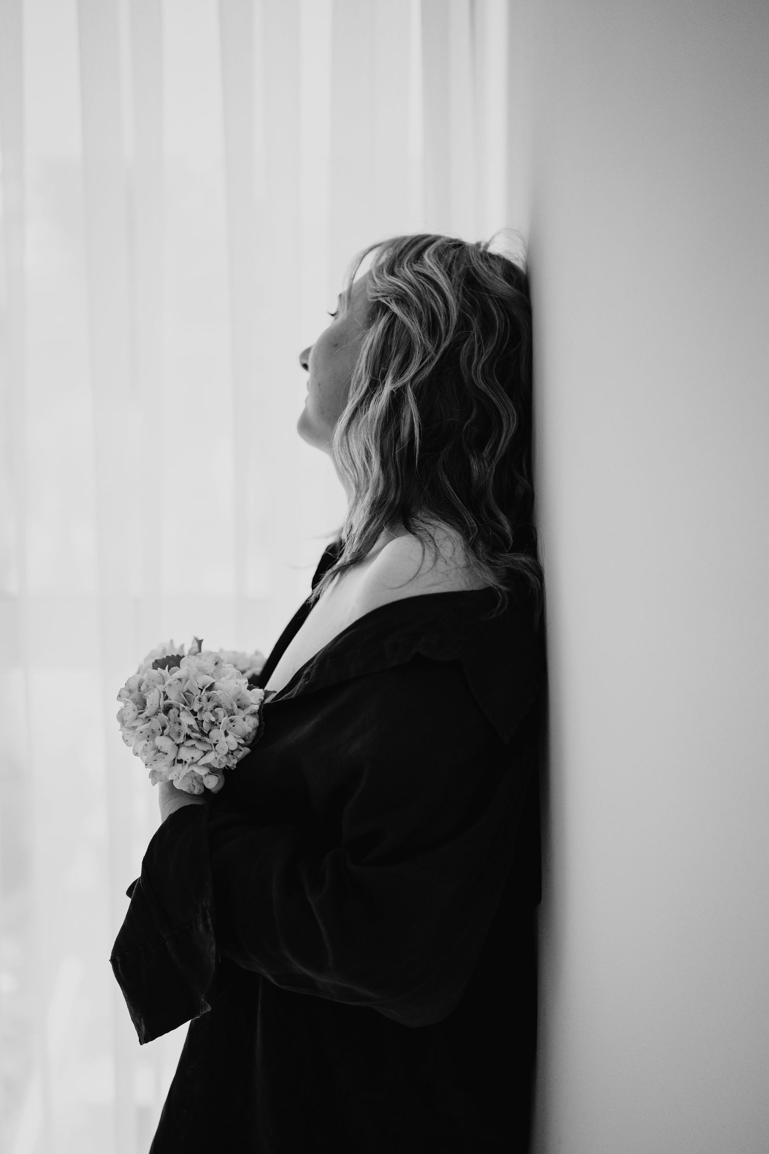 Black and white portrait showing silhouette of figure with flowers in dramatic side lighting against white wall.