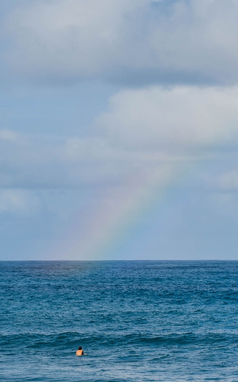 Rainbow appears over deep blue ocean waters with small figure visible in foreground.