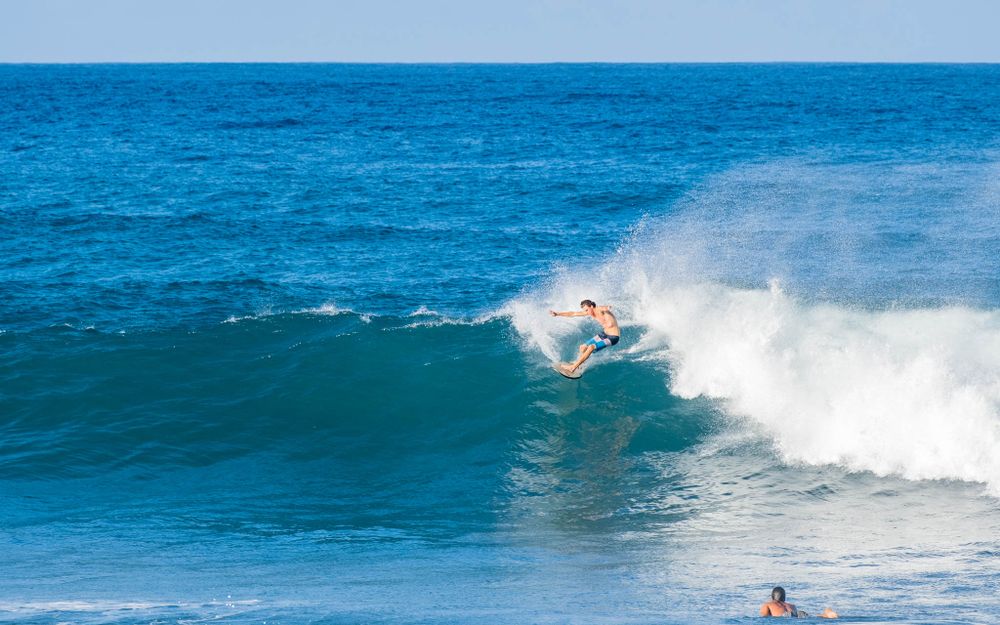 A surfer rides the lip of a clean blue wave while performing a cutback maneuver.