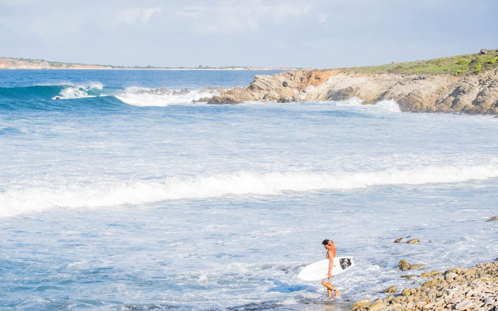 A lone figure stands at the rocky shoreline watching waves break against a coastal landscape.