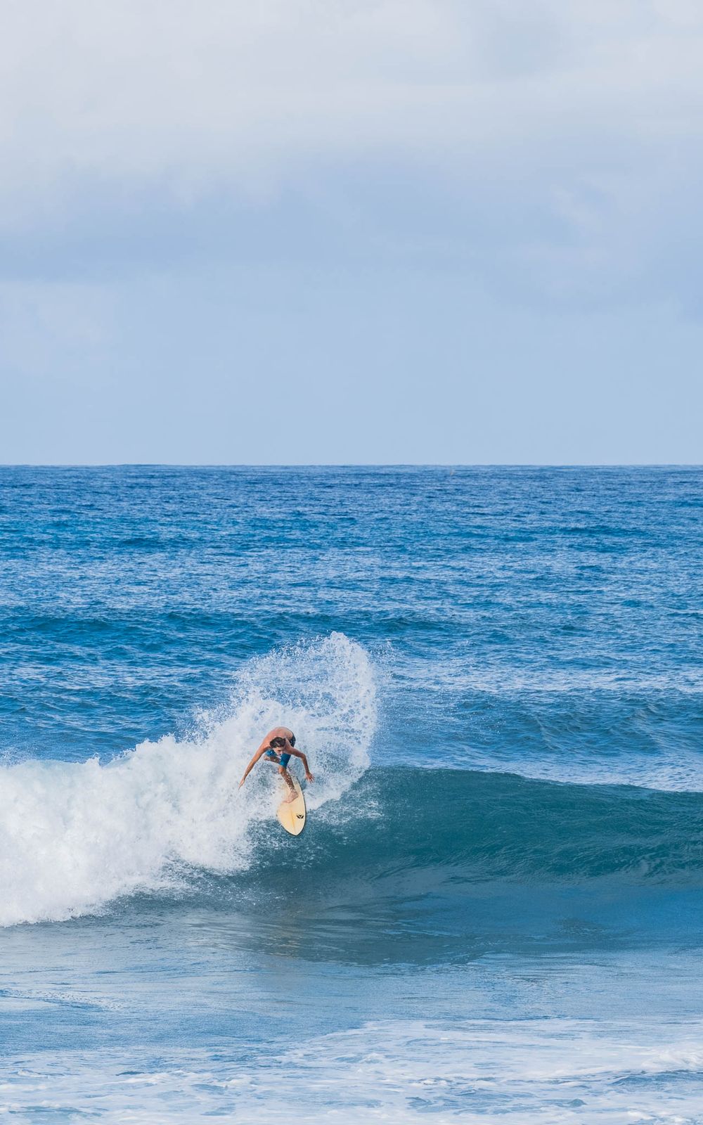 A surfer performs a dynamic turn on the face of a breaking ocean wave against a light blue sky.