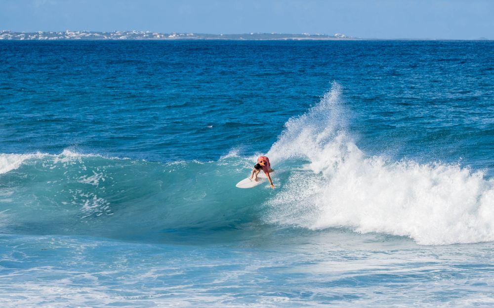 Surfer carves through a breaking turquoise wave with white spray against distant cityscape.