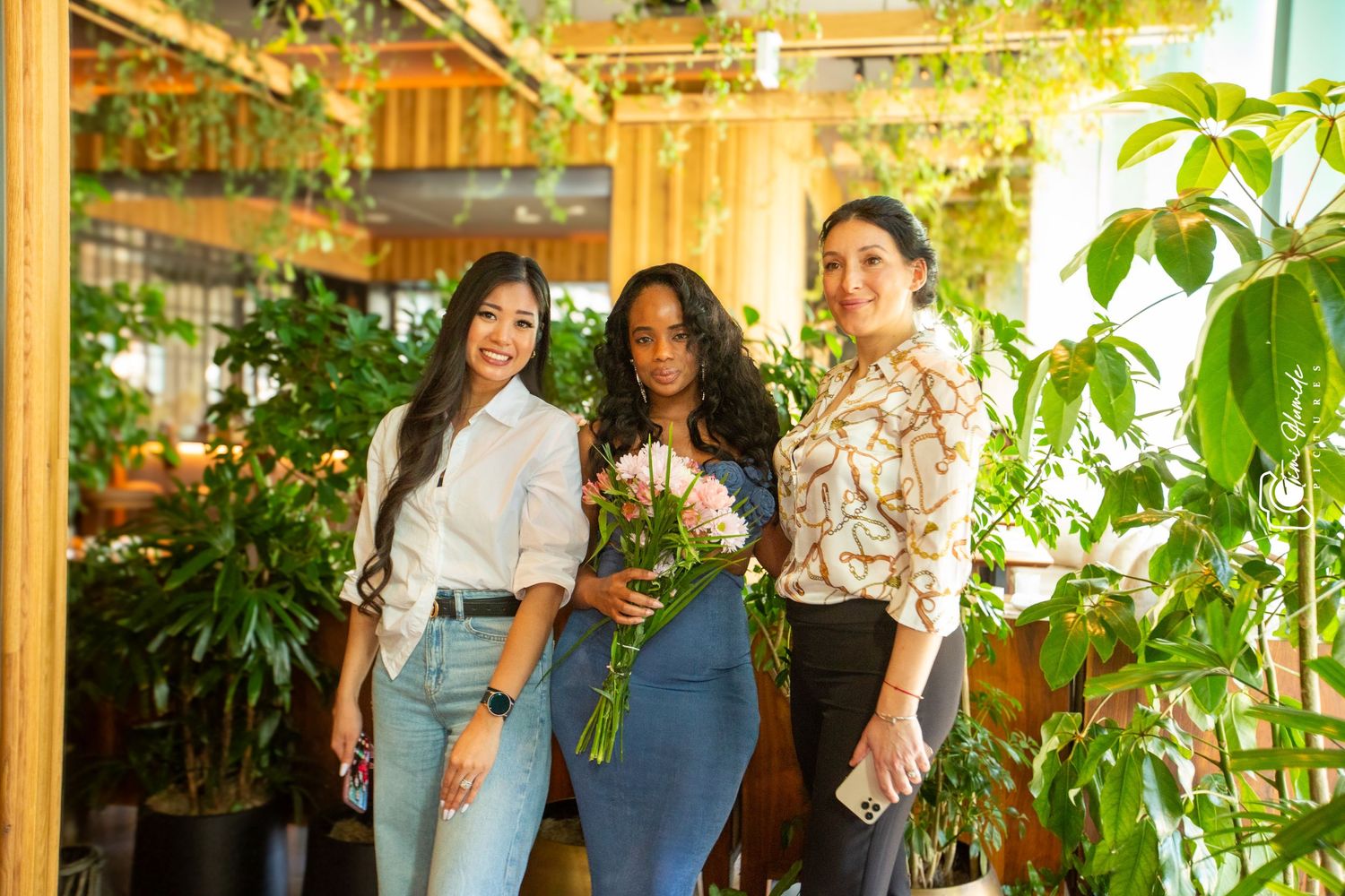 Three women pose casually in a lush indoor garden setting with hanging plants.