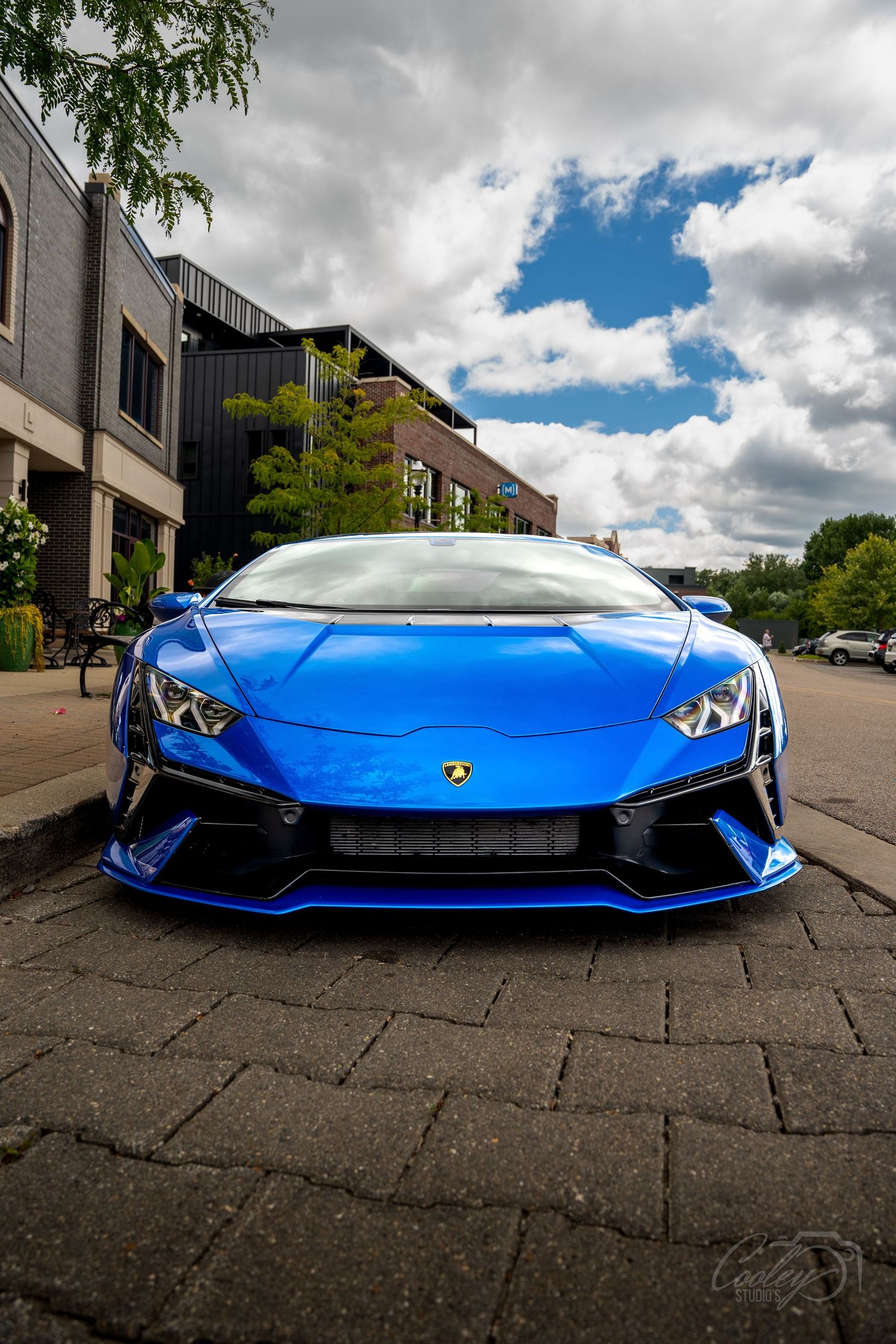 Vibrant blue Lamborghini Huracan parked on brick pavement under dramatic cloudy sky.