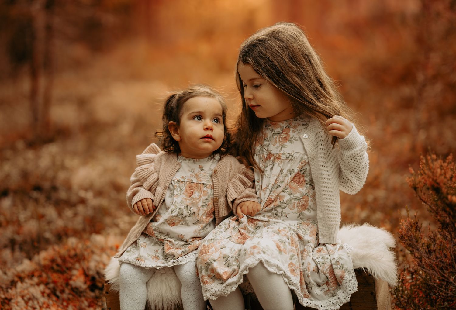 Two children in matching floral dresses sit together against an autumn backdrop of orange foliage.