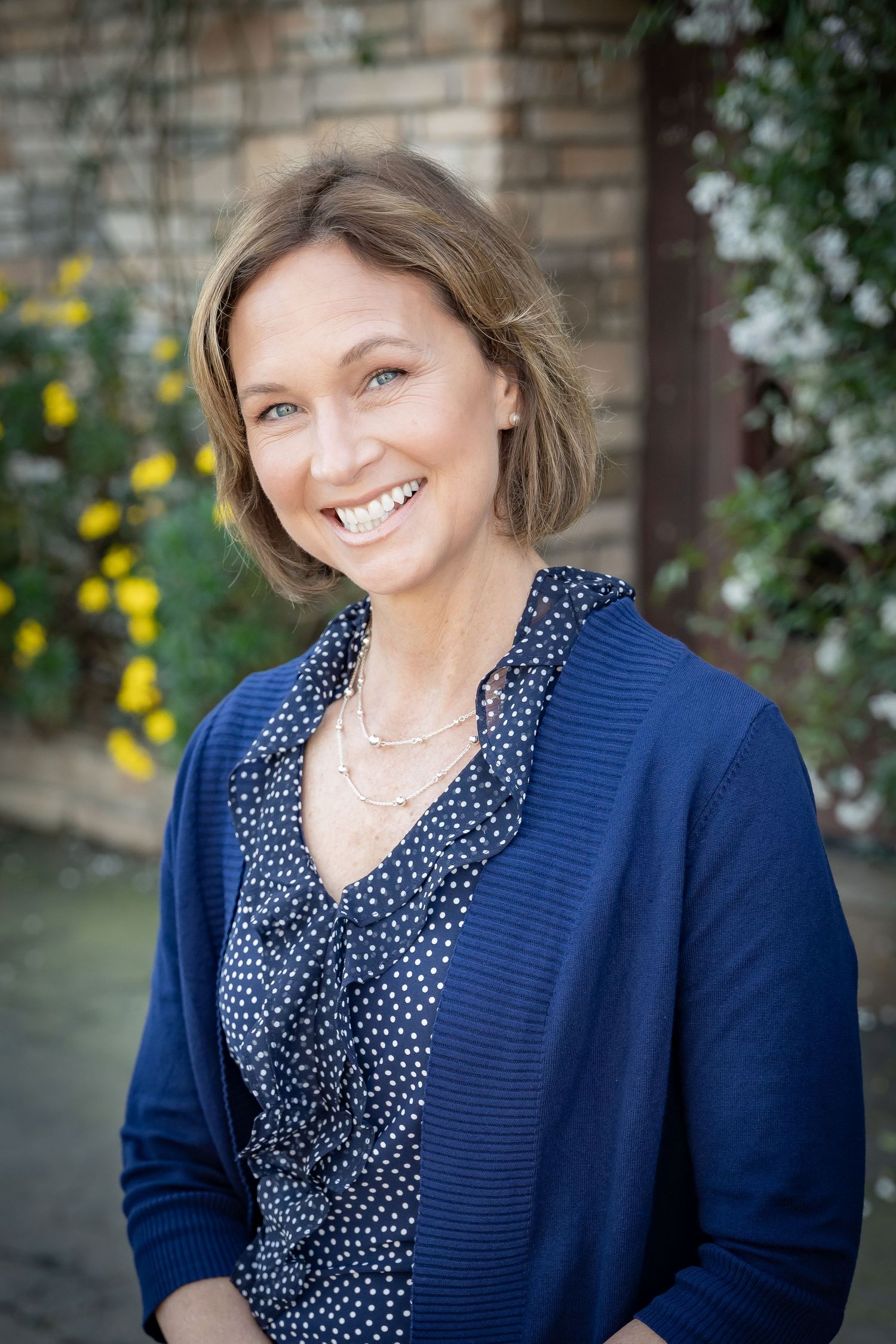 Professional headshot against a brick wall with navy top, polka dot blouse, and garden greenery in background.