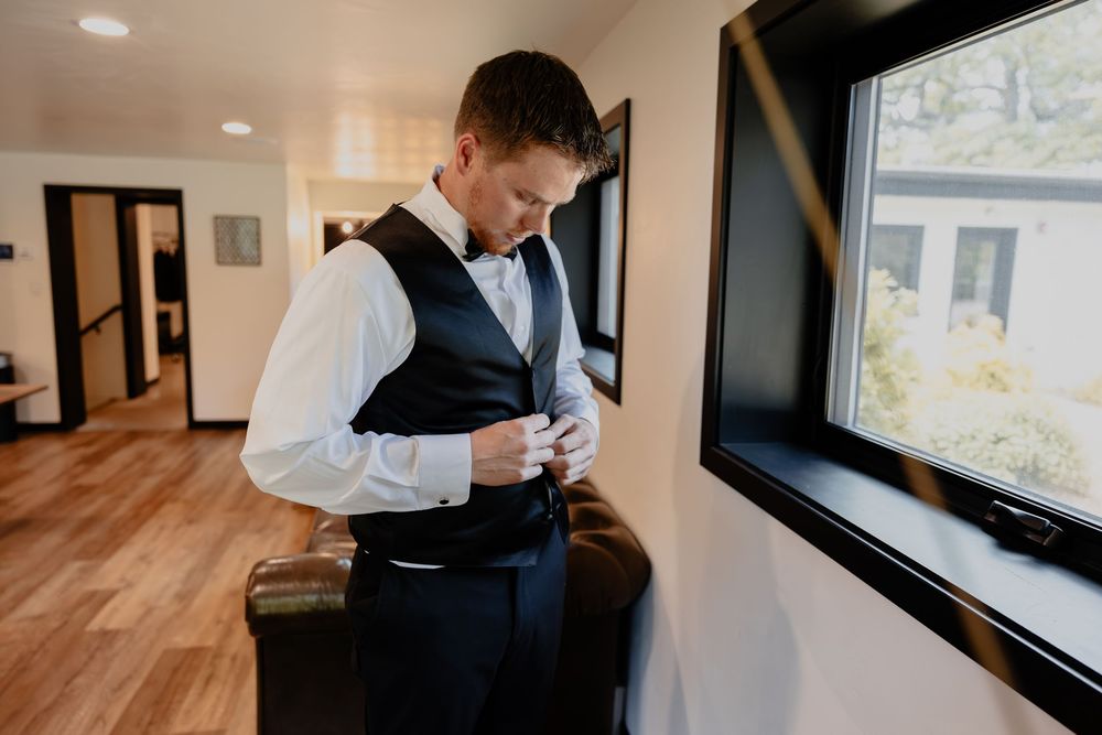 Groom in white shirt and navy vest buttoning up while getting ready indoors on wedding day.